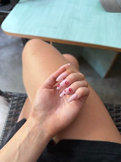 A person's hand with light pink, almond-shaped nails adorned with small red floral accents rests on their thigh. The background shows a light blue table with a light wood trim, a wicker chair, and what appears to be a paved area. The scene is set in Prosena, Bulgaria.