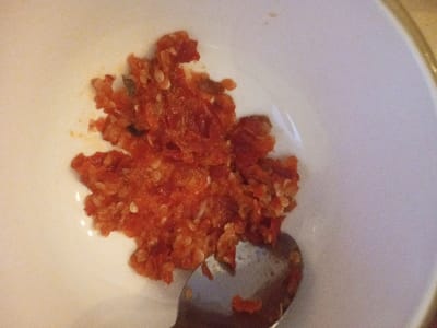 A close-up, overhead view depicts a quantity of coarse, bright red paste, consistent with a chili condiment, situated within a white ceramic bowl. Numerous small, pale seeds and some darker flecks are embedded in the paste, which exhibits an uneven texture. The bowl's upper right edge shows a subtle beige rim. A silver-colored metal spoon, with its bowl submerged in the paste, is visible at the bottom center. The scene occurs indoors in the city of Kanpur, India.