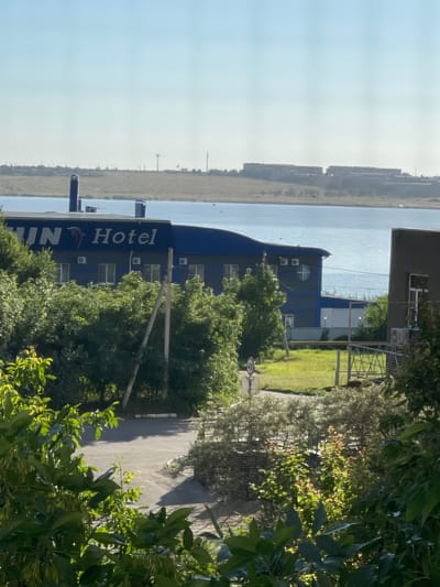 A wide shot captures a hotel building beside a body of water under a clear sky. The hotel, with a blue roof and gray walls, has the word "Hotel" in large white letters on its facade, along with a dolphin logo. Several windows and air conditioning units are visible on the building. In the foreground, lush green trees and bushes partially obscure the view. Beyond the hotel, the water stretches towards a distant, hazy shoreline with buildings and communication towers. A paved area with a speed limit sign indicating "40" is visible near the hotel. The scene appears to be located in Pokoinoe, Russia.
