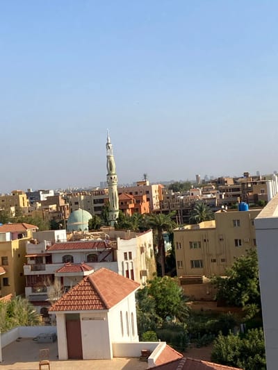 The image depicts a rooftop view of the city of Omdurman, Sudan, with a clear blue sky. The view is from a building located at 15.683 latitude, 32.501 longitude. There are numerous buildings in the scene, with a mosque, a tall minaret, and multiple residential buildings. The buildings are made of brick and concrete, with tiled roofs. There are numerous palm trees and other vegetation growing around the buildings. Some of the buildings are in a state of disrepair, with peeling paint and cracked walls. The image was captured during the daytime, likely midday, as the shadows are not very long. There are no people present in the image, only the buildings and vegetation.