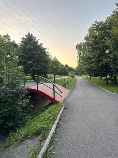 A paved pathway curves through a park in Oktyabr'skoe, Russia. Trees line the left side of the path, with a small red bridge arching over a creek to the right. The bridge has green railings. The time of day appears to be late afternoon or early evening, with a soft, warm light casting a glow on the scene. The sky is a pale yellow and gray, suggesting the sun is low on the horizon. Power lines are visible stretching across the sky in the background. The weather seems calm and pleasant. The scene evokes a sense of peacefulness and tranquility. There is no visible text in the image.