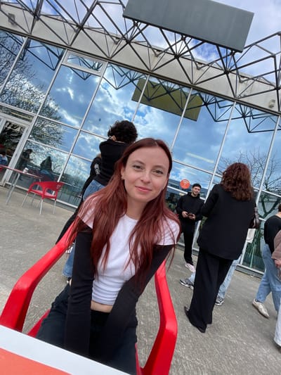 The image depicts an outdoor scene in Bron, France, featuring a group of individuals near a modern building with a reflective glass facade. In the immediate foreground, a female with long, reddish-brown hair is seated in a red plastic chair, facing the camera with a slight smile. She wears a white short-sleeved shirt over a black long-sleeved shirt and dark pants. To her left and slightly behind, another individual with dark, curly hair in a black top is partially visible. Further back, a male in a black hoodie and dark pants stands, looking right while holding a smartphone. To the female's right, a person with dark, curly hair is viewed from behind, wearing a black blazer and wide-leg black pants. Other indistinct figures are visible further in the background, both inside and outside the building. The building features a grey structural framework above its extensive glass panels, which reflect a partly cloudy blue sky and bare tree branches. An empty rectangular sign structure is present at the building's top. Red plastic chairs and small tables are arranged on the paved outdoor area, and a red/orange circular logo is visible on a glass panel.
