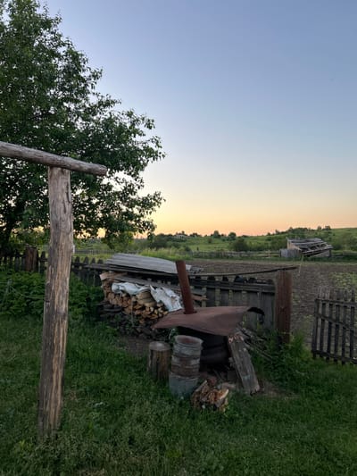 The image depicts a rural outdoor scene at dusk, likely in the village of Борисово, Russia. In the foreground, a weathered wooden post stands to the left, supporting a horizontal beam. Adjacent to the post is a pile of firewood covered with a white tarp. To the right of the firewood is a rusty, barrel-shaped metal structure with a chimney pipe, possibly a makeshift stove or boiler, surrounded by buckets and chopped wood. A dilapidated wooden fence runs horizontally across the midground, separating the immediate area from a tilled field. Beyond the field, a damaged wooden structure with a collapsing roof is visible, and rolling green hills with scattered trees and distant houses stretch towards the horizon. The sky is a clear gradient of pale blue and soft orange, indicative of sunset. The ground is covered in grass and some weeds. No people are present in the scene.