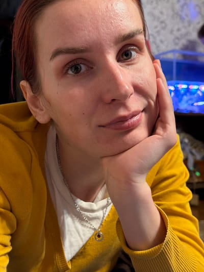 This is an angled, close-up portrait of a person, likely a woman, set indoors in what appears to be a home in Заболотово, Russia. The subject is positioned diagonally across the frame, looking directly at the camera.
The primary subject is a woman with fair skin, light-colored eyes (appearing hazel or grayish-green), and reddish-brown hair. Her eyebrows are dark and distinct. Her lips are a natural pink color, and her skin shows natural texture with some minor blemishes. She has a neutral to slightly pensive expression, with her eyes open and engaged.
She is wearing a mustard yellow, ribbed knit cardigan over a plain white t-shirt. A thin silver chain necklace with a small, simple circular pendant hangs around her neck. Her left hand is resting against her face, with her chin cradled, suggesting a relaxed pose or a moment of contemplation.
The background is out of focus but reveals blurs of bright, colorful artificial lights, possibly from an electronic device, an aquarium, or decorative lighting, consistent with a domestic setting. The image lacks specific cues for the time of day, but the artificial lighting in the background hints at evening or low light conditions indoors. There is no visible text in the media file.
