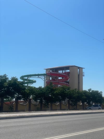 A water park attraction with red and green slides is visible against a clear blue sky in Ömirzaq, Kazakhstan. The slides are part of a tower structure with a covered roof. In the foreground, a row of green trees lines a yellow-walled fence. A paved road and sidewalk are in front of the fence, and a silver car is parked on the sidewalk behind the trees. The scene appears to be an outdoor daytime setting.