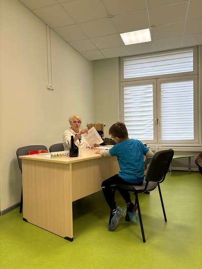 In a brightly lit room with pale green walls and a light beige ceiling, two individuals are interacting at a wooden desk. The room features a large window with vertical blinds and a fluorescent light fixture in the ceiling.
The primary subject is a young boy, appearing to be a student, wearing a blue long-sleeved shirt and black pants. He is seated on a black chair, facing away from the camera, leaning over the desk and looking at a document. He has on blue shoe covers. Next to him, seated on a similar black chair, is an adult woman. She has blonde hair and is wearing a white top. She is also looking at a document and appears to be speaking or explaining something, gesturing with her right hand. She also has on shoe covers.
The desk is positioned in front of the window and is laden with papers and some stationery items, including what appears to be a stapler and a small calendar with colorful pictures. A red container is partially visible on the desk.
The scene suggests an educational or consultation setting, possibly a school or medical office. The presence of shoe covers might indicate a sterile or clean environment. The overall mood seems focused and instructional. The time of day is not explicitly indicated, but the bright lighting suggests daytime. No weather cues are visible. There is no visible text in the image that is legible.