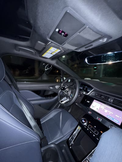 The image captures the interior of a modern car, likely at night or in low-light conditions, as indicated by the illuminated displays and reflections. The view is angled from the passenger side, looking towards the driver's seat and dashboard.

The dashboard features a prominent, large infotainment screen displaying a navigation map alongside a music player interface, showing album art, artist, and song information. Below the screen is a climate control panel with physical buttons and a digital temperature display set to 69 degrees Fahrenheit. Visible controls include fan speed, air conditioning settings, and a red hazard light button. A gear selector, illuminated with a "P" for park, is situated on the center console, which also has a storage or wireless charging area.

A multi-function steering wheel is visible, with various controls and a central logo. The car's interior features dark-colored seats, possibly leather or a synthetic material, with contrasting white stitching. A seatbelt is buckled across the passenger seat. Overhead, the console includes dome lights and controls, some appearing with a magenta hue due to lighting or reflections. A sun visor with warning labels is also visible. Reflections of exterior lights or structures can be seen in the rearview mirror and windshield. Part of a person's leg, clad in blue jeans, is visible on the far right of the frame, suggesting someone is seated in the passenger seat.