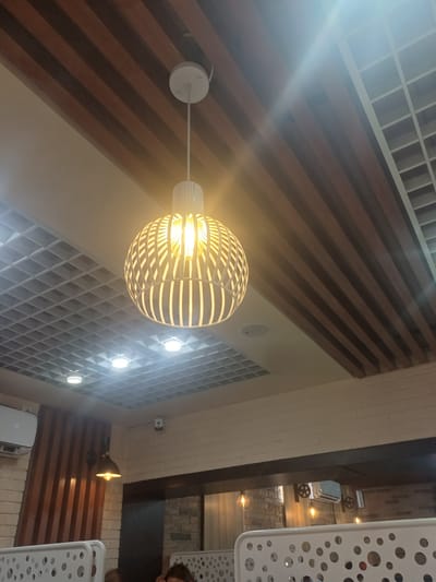 An interior shot of a restaurant ceiling displays a modern pendant light with a white, latticed globe illuminating a warm yellow bulb. The ceiling features a combination of wooden slats and a white grid pattern. Several recessed ceiling lights are visible, some casting bright, circular glows. To the left, a white air conditioning unit is mounted on a brick wall, and to the right, more decorative pendant lights hang in the background. The scene appears to be in the city of Jumashuy, Uzbekistan.