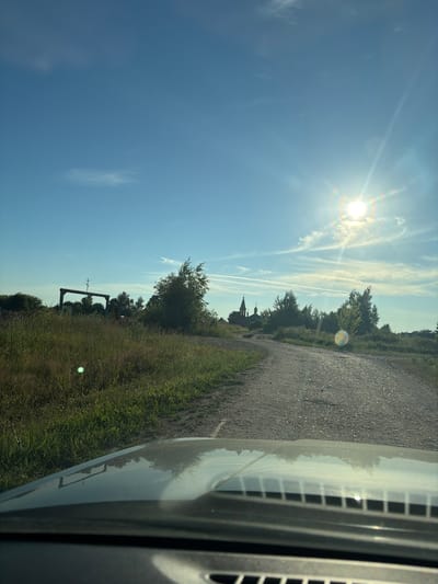 This is a photograph taken from the interior of a vehicle, looking forward and slightly upward. The foreground shows the hood of the car, with a dark, reflective surface and some ventilation grates. The road ahead is a gravel path winding through a grassy, rural landscape. On the left side of the road, a wooden archway with a cross on top stands in a field of tall grass. In the distance, a cluster of trees frames a church with a green dome and a steeple. The sky is a clear blue with a few wispy white clouds and the sun is shining brightly, creating lens flare and some rainbow effects. The scene appears to be set in Pobedinka, Russia, given the distinctive architectural style of the church and the rural setting.