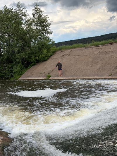 The photograph depicts a waterfall and adjacent concrete structure in Nizhnii Vishur, Russia. A person stands barefoot on the concrete near the waterfall's edge. The setting includes a mix of natural and man-made elements, with trees and sky visible in the background.