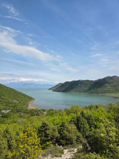 The image shows a view of Skadar Lake in Godinje, Montenegro. The lake is surrounded by green mountains and dense vegetation. The sky is light blue with streaks of clouds. In the distance, mountains are visible through a haze.