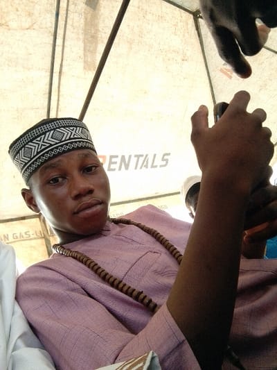 A young man wearing a traditional cap and a light pink shirt with a wooden bead necklace poses for a selfie. He holds a phone in his right hand. The background is a white tent with visible support beams. The image is taken from a low angle, looking up at the man. Another person, wearing a white hat, is partially visible behind the man's arm. Part of a banner with the word "RENTALS" is also visible.