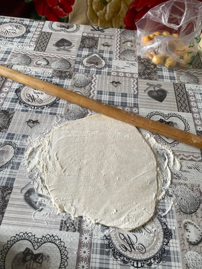 The image shows a flat, uncooked dough with a rolling pin on a table covered with a patterned tablecloth. The tablecloth has a patchwork design with various motifs, including hearts, lace-like patterns, and plaid sections. Flour is scattered around the dough and on the tablecloth. In the upper right corner of the image, a clear plastic bag filled with small, light-colored fruits, possibly apricots or plums, is visible. The background shows a blurred red and yellow fabric, suggesting a domestic setting. The scene appears to be a kitchen or dining area, possibly in Lebedinovka, Kyrgyzstan, where dough is being prepared for cooking.