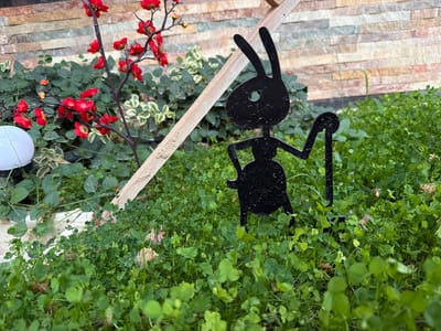 The image depicts a close-up view of a garden area with lush green clover in the foreground. A black silhouette of an ant-shaped garden ornament stands prominently in the center, its antennae and limbs clearly defined. To the left, a decorative branch with small red flowers adds a splash of color. Behind the plants, a stone wall with horizontal bands of various earthy tones forms the backdrop. A light-colored wooden plank cuts diagonally across the upper portion of the image, appearing to be part of a garden structure or support. A white spherical object is partially visible on the far left, possibly a garden light. The scene suggests an outdoor setting, likely a garden or courtyard, with a focus on decorative elements and plants. The lighting suggests it could be daytime, but the specific time or weather conditions are not apparent. There is no visible text in the image.