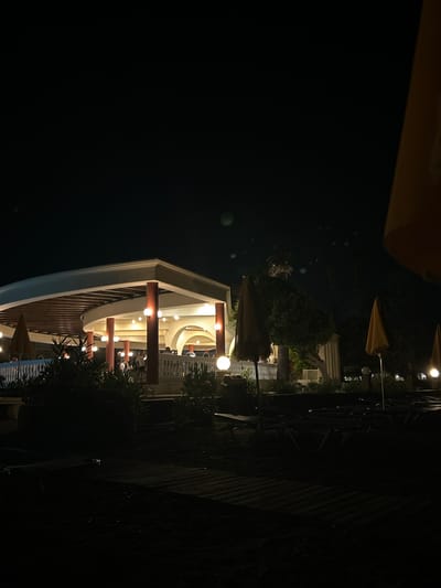 This is a nighttime, low-angle, full-shot photograph of a resort's outdoor dining area and beach chairs on the island of Rhodes, Greece. In the background, a curved, open-air pavilion with a slatted wooden roof is illuminated by numerous warm, spherical lights. The pavilion has white balustrades and is supported by reddish-orange columns. People are visible inside the pavilion, appearing to be dining or socializing. In the foreground, dark silhouettes of lounge chairs and beach umbrellas are arranged on the sand, with a wooden walkway in the very front. Several small, round lights are visible near the ground in front of the pavilion. The sky is dark, with a few indistinct blurry lights, possibly lens flares or atmospheric phenomena. The overall impression is of a tranquil, after-hours resort ambiance.