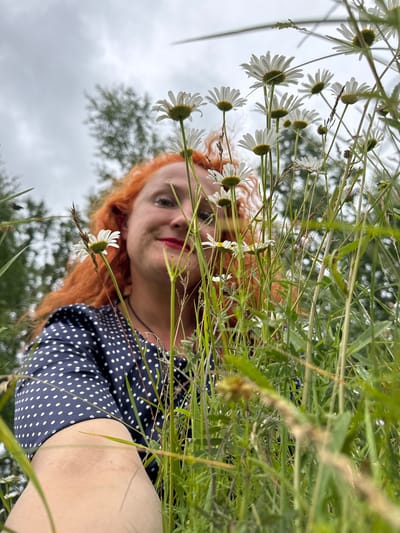 A low-angle shot captures a woman with bright red, curly hair smiling and looking at the camera from behind a cluster of white daisies. She is wearing a dark blue dress with white polka dots. The background is a mixture of green grass, trees, and a cloudy sky. There is no specific indication of the city of Privolzhsky, Russia.