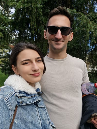 A selfie depicts a young woman and a man posing outdoors in Rheinfelden, Switzerland. The woman has dark hair and is wearing a denim jacket with a white faux fur collar. She is leaning against the man. The man has short dark hair, a light-colored long-sleeved shirt, and sunglasses perched on his nose. Behind them is a dense green evergreen tree. A partially visible water bottle sits on the ground to the lower left. The sky is partially visible through the tree branches. The scene appears to be taken in a park or green space, typical of Rheinfelden's urban landscape.