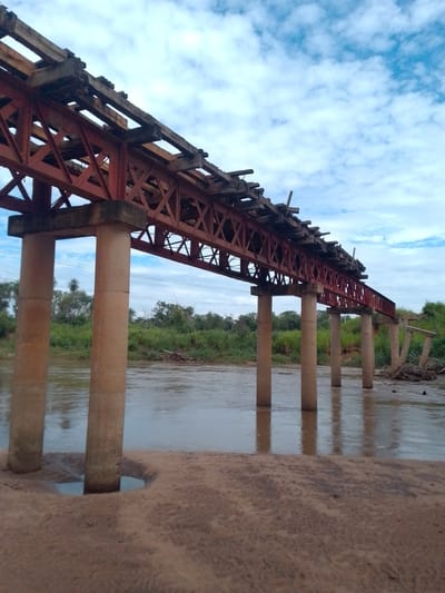 The image depicts a damaged railway bridge spanning a river, likely taken near Horqueta, Paraguay. The bridge structure is primarily steel, painted red, with wooden supports on the upper level. Several support pillars stand in the water, but one section of the bridge has collapsed, with debris visible in the river. The river is wide and calm, reflecting the bridge and the cloudy sky. The riverbank in the foreground is sandy and shows signs of erosion around the bridge supports. Lush green vegetation lines the riverbanks in the background.