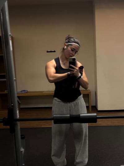 The image depicts a female individual taking a mirror selfie inside an indoor gym facility in Komchén, Mexico. She is centered in the frame, wearing a black sleeveless tank top, grey sweatpants, a white and black patterned headband, round glasses, and white wireless earbuds. Her dark hair is pulled back. She holds a dark-colored smartphone in her right hand, pointing it towards herself, and also clutches a dark-colored water bottle or shaker under the same arm. Her left arm is flexed, showcasing muscle definition. A smartwatch is visible on her left wrist, and multiple bracelets adorn her right wrist. The background features light beige walls, a dark gym floor, and a light-colored wooden bench. To the left, a metal gym rack is visible, with a barbell fitted with a black pad.