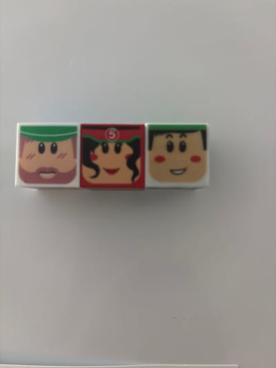 The media file displays three stacked blocks, each featuring a cartoon face. The blocks are oriented vertically. The top block shows a smiling face with black hair and red cheeks, wearing a green headband. The middle block depicts a face with dark hair styled in pigtails and red lips, enclosed in a red frame with a white circle containing the number "5". The bottom block has a face with light brown hair and rosy cheeks, wearing a green cap. The background is a plain, light-colored surface, likely a table or desk, with some subtle smudges. The overall scene suggests a toy or game made of these blocks. The location context of Княгинино, Russia, is provided but not visually evident in the image itself.