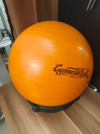 An orange "GymnastikBall" manufactured by Ledragomma in Italy is pictured on a wooden surface. The ball has horizontal ridges and a logo that reads "ORIGINAL PRESS GymnastikBall Made in Italy by LEDRAGOMMA ø max cm. 53". A dark grey base supports the ball. The background includes a dark vertical surface and a lighter wooden surface with visible grain. The lighting suggests an indoor environment. The location is not discernible from the image.