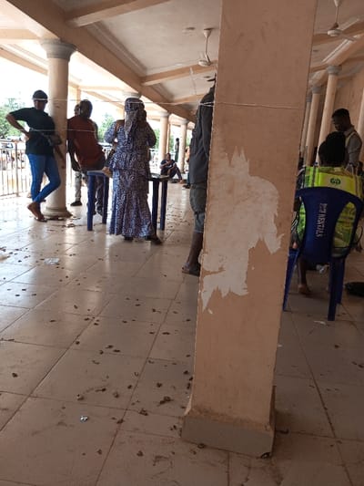 The image displays a semi-enclosed, columned walkway or veranda in Ajalli, Nigeria. The floor is tiled with light-colored square tiles and is heavily littered with numerous small, dark, irregularly shaped items, consistent with nuts or seeds, along with scattered pieces of light-colored debris. Cream-colored columns with grey capitals support an overhead structure containing multiple ceiling fans.
Seven individuals are present. In the left midground, a person wearing a black t-shirt, blue jeans, and sandals leans against a column, hands on hips, with a dark bag across their chest. To their right, another individual in a reddish-brown shirt stands. In the central midground, a person wearing a blue and white patterned long dress and a head covering stands upright, facing away from the viewer, near a small blue plastic rectangular table. A prominent, cream-colored rectangular column in the right foreground shows significant paint peeling, revealing a lighter surface underneath. Behind this column, a person wearing a bright green and yellow reflective vest over a dark shirt is seated on a blue plastic chair, the back of which reads "RENTALS." Further in the background, a person in a light-colored shirt stands, and another individual is seated further back. A thin, light-colored string or wire is stretched horizontally across the midground between columns. The scene appears to be an active, public gathering or waiting area under natural daylight.
