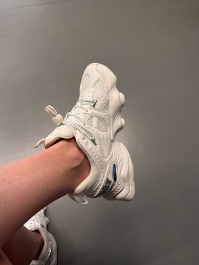 A close-up, eye-level shot shows a person's left leg and foot wearing a white, chunky-soled athletic shoe against a plain gray background. The shoe has a mesh upper, white laces with a toggle closure, and metallic blue accents on the side. The leg appears pale and hairless, and the ankle is visible as it enters the shoe. The lighting is soft and even, creating no harsh shadows. The image is framed with the shoe in the foreground, taking up the majority of the right side of the frame, and the leg extending from the bottom left corner. No identifying features of the person are visible. There is no indication of the location being Kyrenia, Northern Cyprus or Cyprus; the background is a nondescript gray surface.