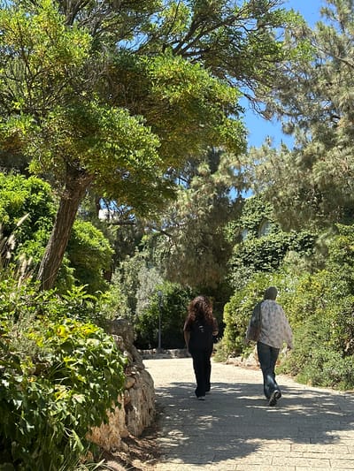 Two individuals walk away from the viewer on a paved path in a lush, green outdoor environment. The person on the left has long, curly brown hair and wears dark clothing and a black backpack. The person on the right wears a headscarf, a light-colored floral shirt, and blue jeans, and carries a bag over their shoulder. The path is lined with stone on the left and abundant vegetation, including trees and bushes, on both sides. In the background, a building is partially obscured by dense green foliage. The scene appears to be a park or garden area. The image location is identified as the city of al-Eizariya, Palestinian Territories.