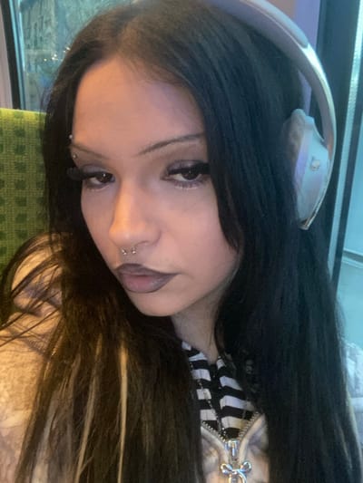 A head-and-shoulders portrait depicts a female subject seated inside a public transport vehicle in Enghien-les-Bains, France. The subject occupies the majority of the frame, positioned slightly off-center to the left. She has long, dark hair with a visible light-colored streak on the viewer's right side, parted down the middle. She wears light-colored, over-ear headphones resting on her head and right ear. Her face features notable makeup, including dark lipstick, prominent winged eyeliner, false eyelashes, and white eyeliner on the lower lash line. Facial piercings are evident: a three-stud eyebrow piercing above her right eyebrow and a silver-toned horseshoe-shaped septum ring. She wears a black and white striped top under a partially unzipped light-colored hoodie, accessorized with a silver-toned chain necklace featuring a stylized bow-shaped pendant. The background consists of a blurred interior, with a green textured surface (likely a seat) to the viewer's right and a window to the viewer's left showing faint, cool-toned urban or suburban lights, suggesting an evening setting.