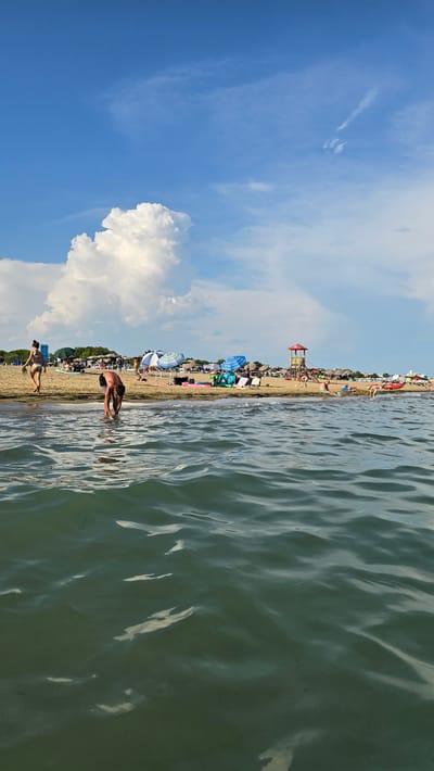The image depicts a vibrant summer day at a crowded beach in San Michele al Tagliamento, Italy. The scene is dominated by the juxtaposition of a bright blue sky filled with fluffy white clouds and the tranquil, rippling surface of the sea. The beach itself is a hive of activity, with numerous sunbathers scattered across the sand, many under colorful umbrellas, enjoying the warm weather.

In the foreground, two individuals are visible near the water's edge. One person, a man, is captured in a dynamic pose, bending down near the shallow water. Further along the shore, a woman in a bikini is walking away from the camera. The presence of beach chairs and umbrellas suggests a well-established recreational area. A lifeguard tower, painted in red and white, stands prominently on the beach, indicating safety measures are in place.

The water is a dominant feature, its surface reflecting the sunlight and creating a shimmering effect. The background shows a dense line of trees behind the beach, hinting at the coastal vegetation. The overall impression is one of a lively and enjoyable summer day, with people relaxing, swimming, and engaging in beach activities. The bright sunshine and clear skies suggest it is midday or early afternoon. No visible text is present in the image.