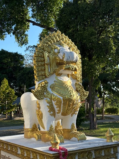 This image features a white statue adorned with gold embellishments, likely a mythical creature or guardian, common in Buddhist art. The statue has a detailed, ornate headpiece and intricate gold patterns across its body, including what appears to be wings and a textured mane. It stands on a white platform with gold trim.

The setting is outdoors, with lush green trees and bright blue sky visible in the background. The lighting suggests it's daytime, likely with sunny weather given the clear sky and defined shadows. There are indications of a tropical environment, with the types of trees and the overall atmosphere. The statue's presence suggests a religious or cultural site, possibly a temple or shrine, with the location context pointing to Baan Tha Po, Thailand. No people are visible in the frame. The focus is entirely on the statue and its surroundings. The statue itself conveys a sense of majesty and perhaps protection, typical of such artistic representations. Some text might be present on the gold trim of the platform, but it is not clearly legible in this image.