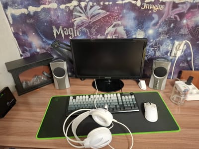 The image depicts a desk setup in a room. In the foreground, a wooden desk is visible, holding various computer accessories. A black mousepad with a green border is present, on which a white computer mouse and a mechanical keyboard are placed. A pair of white headphones rests in front of the keyboard. To the left of the keyboard, a black router with green indicator lights is situated. Behind the keyboard and mousepad, a black computer monitor is centered on the desk. To the left of the monitor, a small, dark grey speaker is positioned, and to its right, another identical speaker is placed. A decorative item resembling a miniature fireplace with orange and red elements is visible to the far left of the desk. The background features a wall adorned with a tapestry depicting a celestial scene with stars, a nebula, a book, and a ship, along with text that reads "Magic" and "Imagine." A power strip with multiple outlets and plugs is mounted on the wall to the right of the monitor. The overall environment appears to be a personal workspace. The image does not provide any information that would allow for the determination of its location in the city of Novlyanka, Russia.