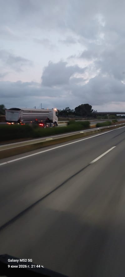 A white dump truck is traveling on a highway in Vila-seca, Spain. The truck is carrying a load of dark material and has its headlights and brake lights on. The highway is paved and has white lines marking the lanes. There are trees and vegetation on either side of the highway. The sky is cloudy and appears to be dusk or early evening. The text overlay indicates the image was taken on June 9, 2026, at 21:44.