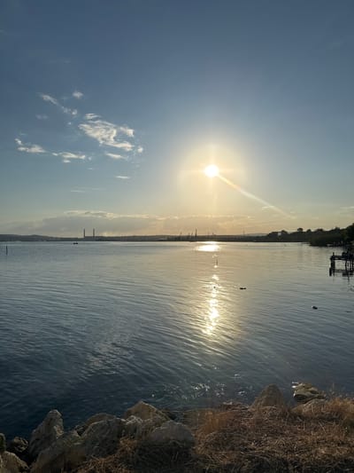 The image captures a serene coastal view at sunset in the city of Kazashko, Bulgaria. The sun, positioned in the upper center of the frame, casts a warm, golden glow across the rippling surface of the water, creating a shimmering reflection. Scattered clouds drift across the expansive blue sky. In the distance, a hazy industrial landscape is visible along the horizon, characterized by tall structures and cranes, suggesting a port or manufacturing area. The foreground features a rocky shoreline with dry grass and vegetation, leading down to the water's edge. There are no people visible in the scene. The overall atmosphere is peaceful and tranquil, dominated by natural light and the vastness of the sky and sea.