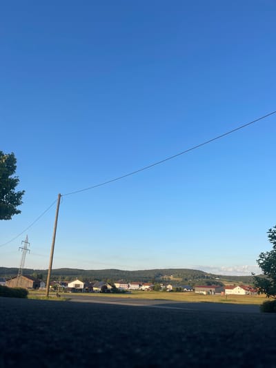 The image shows a rural scene in Cham, Germany, with a clear blue sky. In the foreground, a dark, textured wall runs along the left side of the frame. A paved road is visible next to the wall, leading into a landscape dotted with houses and trees. A wooden pole stands upright, with a wire extending from its top towards the right side of the image, disappearing into the vast blue sky. In the distance, a gentle slope covered in trees creates a backdrop to the scattered buildings. A power line tower is also visible in the middle ground, indicating the presence of electricity infrastructure. The sunlight suggests it is daytime, likely morning or afternoon, given the clear and bright sky. The scene evokes a sense of peace and rural tranquility.