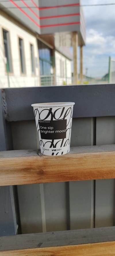 A disposable white paper coffee cup is positioned on a weathered wooden ledge against a gray metal structure. The cup is printed with a black and white design featuring stylized graphics and text that reads "#good mood" and "One sip brighter mood." The branding "DOLCE Vending market" is printed at the bottom of the cup.

The background includes a blurred building facade, with white walls, dark-framed windows, and a roofline trimmed with red accents. Faintly visible beyond the building is a cloudy sky.

Based on the cup branding "DOLCE," a company based in Hovtashen, Armenia, it can be assumed the photograph was taken in this location.