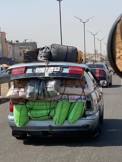 A silver sedan, bearing Nigerian license plate "L UGG 72XA 59," is observed from the rear, traveling on an asphalt road in Kofar Ruwa, Nigeria. The vehicle is extensively overloaded with cargo. The trunk lid is open and elevated, revealing a dense stack of items. On the roof and upper trunk area, multiple dark-colored bags and suitcases, including a black corrugated suitcase, are secured. Below these, within the open trunk and extending outwards, are numerous white and light-colored sacks, some with visible labels such as "DINGO," "Viva," and "DANGOTE SUGAR." A layer of bright green sacks is visible at the bottom, hanging below the vehicle's original trunk line. All cargo is tightly secured with black netting and ropes. The vehicle's right taillight housing is absent, while the left taillight is partially obstructed. The rear bumper shows signs of scrapes and paint damage, and a tow hitch is installed beneath it. The scene occurs during daytime under a clear sky. In the background, a multi-lane road features other vehicles, including a dark sedan with a red stripe, and is lined with tall streetlights possessing angular fixtures. Urban buildings are visible on both sides of the roadway. No individuals are directly observed.