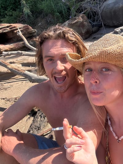A close-up photograph depicts two individuals on a sandy beach in Coalcliff, Australia. The male, positioned on the left, is shirtless with light brown, shoulder-length hair and tan skin. He wears blue shorts and looks towards the camera with an open-mouthed expression. The female, positioned to the right and partially visible, wears a straw hat, a patterned swim top, and a light-colored beaded necklace. Her left hand, adorned with a silver ring on the ring finger, holds a cigarette with a silver filter holder between the index and middle fingers. She looks directly at the camera with a neutral expression. The immediate background shows sandy ground with scattered driftwood and large rock formations. Further back, green foliage is visible on an elevated landform. The scene is illuminated by bright daylight.