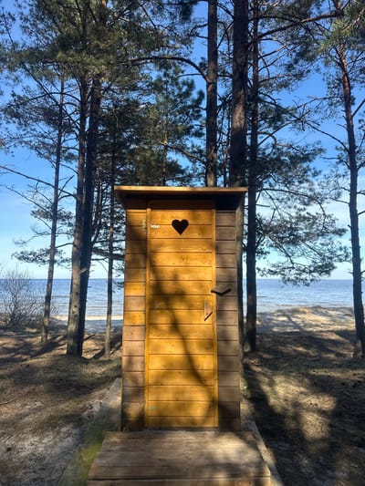 An upright, sunny outdoor scene depicts a rustic wooden outhouse situated among pine trees near a body of water. The outhouse, made of light-colored wooden planks, stands prominently in the foreground, angled slightly to the right. Its door features a distinctive heart-shaped cutout near the top left corner, and a simple wooden latch is visible on the right side of the door. Long shadows from unseen objects or the trees diagonally stripe the outhouse's wooden facade, indicating direct sunlight. A wooden platform or path extends from the front of the outhouse, curving towards the right foreground.
Tall, slender pine trees frame the outhouse and the background, with their dark trunks and green needles contrasting against a clear, bright blue sky. Beyond the trees, a shimmering body of water, likely the Baltic Sea, stretches to the horizon. A sandy or grassy bank with sparse vegetation and pronounced tree shadows descends towards the water's edge.
The lighting suggests a bright day, possibly morning or late afternoon, with clear skies. The scene conveys a peaceful, natural setting characteristic of a coastal forest. No people or visible text are present. The image was captured in Zīvartiņš, Latvia, a location consistent with a coastal, forested landscape.