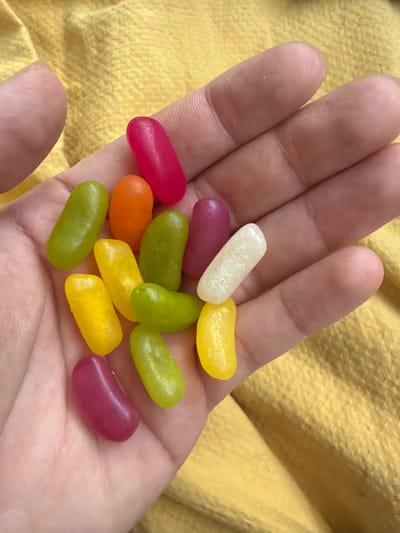 A person's open hand holds an assortment of approximately 12 jelly beans. The jelly beans are various colors: white, red, orange, green, yellow, and purple. The hand is positioned against a textured yellow background. The setting is unknown, and there is no discernible connection to Pentre, United Kingdom.