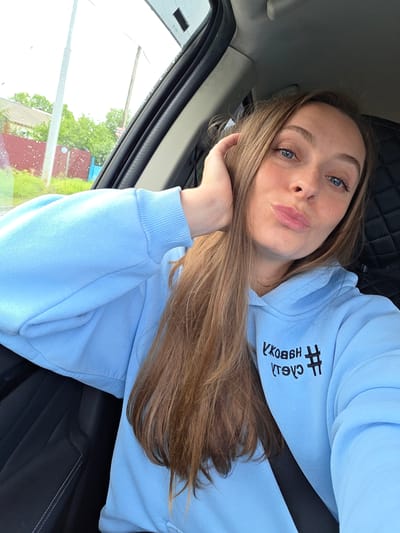 The image depicts a young adult female seated inside a vehicle, consistent with the passenger seat, at a location identified as Valdgeym, Russia. She has long, light brown hair, blue eyes, a light complexion with visible freckles, and is making a pouting facial expression towards the camera. Her left hand is raised, with fingers resting near her ear and hairline. She is wearing a light blue hooded sweatshirt (hoodie) with black embroidered text on the front, which appears mirrored due to the selfie perspective. The text, when read correctly, states '#ЛЮБОВЬ В СЕРДЦЕ' (Love in Heart). A black seatbelt is secured across her chest and shoulder. The vehicle's interior features dark-colored upholstery, including a quilted pattern visible on a seat or panel behind her. Through the rain-speckled car window to her left, an overcast outdoor scene is visible, featuring green trees and grass, a red-painted building partially obscured by foliage, a utility pole, and a distant blue structure, all under a grey sky.