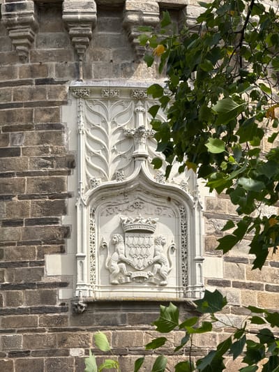 The image shows a close-up, eye-level view of a stone facade on the exterior of a building. The facade is constructed of rough-hewn, rectangular stones in varying shades of brown and gray, laid in a horizontal pattern. There are decorative elements made of lighter-colored stone, including a large, ornate carving in the center. This carving features a shield held by two lions, surmounted by a crown, and framed by intricate foliate designs and architectural elements. The carving is likely a coat of arms or a heraldic symbol. To the right and upper part of the image, a tree with lush green leaves partially obscures the stone structure. The lighting suggests it is daytime, with shadows indicating sunlight.
This facade is part of the Château des Ducs de Bretagne (Castle of the Dukes of Brittany) located in Nantes, France. Specifically, this view is of the Renaissance wing of the castle, which houses a museum. The decorative carving visible is a significant architectural feature of this historic building.