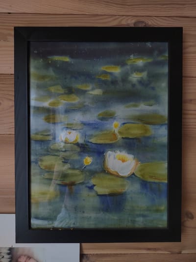 A framed watercolor painting of water lilies is displayed on a wooden wall. The painting depicts several lily pads floating on water, with two prominent white water lilies in bloom. The background is a dark, moody blue-green, suggesting a pond or lake. The frame is simple and black. The wooden wall is composed of horizontal planks with visible grain and knots. The overall scene is calm and serene. The painting is located in the rural area of Бярэзінскі сельскі Савет, Belarus.