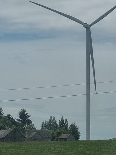 The image shows a close-up view of a wind turbine with its three blades extending upwards. The sky is overcast and a pale blue, with some wispy clouds. The turbine is positioned in an open field, with trees and several rustic-looking buildings visible in the background. The buildings appear to be made of wood and have steeply pitched roofs. A strip of green grass is visible on the right side of the image. The image is oriented vertically, so the scene is presented on its side. The overall impression is one of a rural landscape, possibly on a slightly windy day, given the presence of the wind turbine. There is no text visible in the image. The lighting suggests it is daytime, but the overcast sky gives it a muted appearance. The scene appears peaceful and serene.