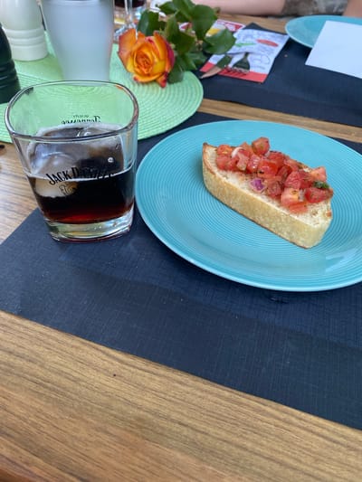 The image captures a close-up view of a table setting, likely at an outdoor dining establishment in Wölfersheim, Germany. The foreground features a glass of dark liquid with ice, branded with "Jack Daniel's". Next to it, on a teal-colored plate, is a slice of toasted bread topped with diced tomatoes and red onions, resembling bruschetta. A decorative orange and yellow rose is visible in the background, resting on a green placemat, alongside what appears to be a white ceramic jug and a darker placemat underneath. The table itself is made of wood. The overall scene suggests a relaxed meal or snack.