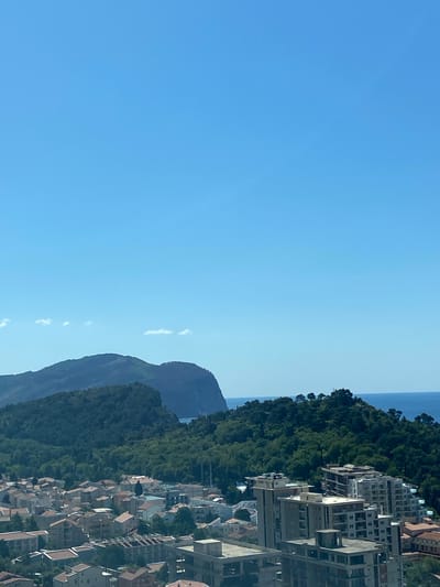 The image presents a panoramic daytime view of a coastal landscape, consistent with the location context of Nerin, Montenegro. In the foreground and mid-ground, a dense cluster of urban buildings, likely residential and commercial, features predominantly light-colored facades and numerous terracotta-tiled roofs. These structures are built along a sloping terrain, suggesting a hillside settlement.
A prominent, densely forested mountain or large hill forms a natural barrier, extending horizontally across the mid-section of the frame, positioned behind the urban area. Beyond this forested expanse, the serene blue waters of the sea are visible on the horizon to the far right. Further to the left and receding into the distance, another, larger and more rugged mountain range with sparse vegetation can be seen, its peaks softened by atmospheric haze.
The sky is a clear, bright blue, indicating sunny weather and good visibility. The strong, even light suggests the time of day is either morning or early afternoon. No individuals, specific activities, or visible text are present in the scene, which conveys a sense of peaceful natural beauty intertwined with human habitation.