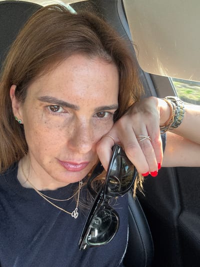 The image is a close-up shot of a woman in what appears to be a car. She is positioned with her head tilted, looking directly at the camera. The woman has light brown hair, freckles, and brown eyes. She is wearing a gold necklace and earrings, as well as several rings on her fingers. Her fingernails are painted red. She is holding a pair of black sunglasses. The background shows a car interior with the sun visor and a glimpse of foliage outside the window. It is not possible to confirm the location in Albergaria-a-Velha, Portugal, from this image alone.
