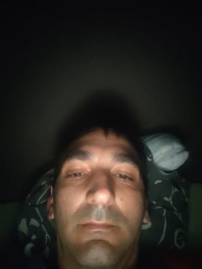 The image depicts a close-up, low-light portrait of a single adult male, presumably taken in the city of Servas, France. The male, appearing middle-aged, is positioned centrally, facing directly towards the camera with a neutral expression. He has short dark hair, distinct dark eyebrows, and visible stubble on his upper lip and chin. His eyes are open and directed forward. Behind his head, to the right, a dark pillow with light grey or white abstract patterns is visible, alongside a lighter, solid grey or white pillow portion. The immediate environment is dark, suggesting an indoor setting with minimal illumination, likely from the device capturing the image, which casts a faint shadow upwards and behind his head against a uniformly dark background. There are no other individuals, subjects, or interactions depicted.