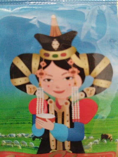 This is a close-up, cartoon-style illustration of a person in traditional clothing. The subject is a young girl, depicted with rosy cheeks, a slight smile, and large, dark eyes. She wears a tall, conical hat with a red and yellow top. Her attire includes a black garment with gold trim, a red circular detail on her shoulder, and blue sleeves. She also has elaborate hair accessories with beads and golden elements. In her hands, she holds a white bowl with red trim.

The background features a bright blue sky with faint white shapes that could represent clouds or mountains, and a green grassy field below. In the distance, a herd of sheep are grazing, and a single brown animal, possibly a cow or horse, is also visible. The overall scene suggests a rural or pastoral setting, likely a meadow or steppe.

The illustration has a cheerful and innocent feel, with no specific time of day or weather indicated beyond the clear sky. There is no visible text in the image. The style is simplified and colorful, characteristic of children's book illustrations or decorative art. The clothing suggests a cultural context, and the inclusion of sheep in a grassy landscape might imply a nomadic or agricultural lifestyle.