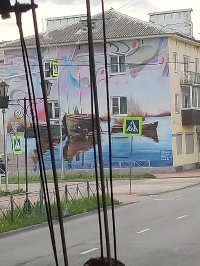 This detailed image captures a two-story building located at Mineralnaya Street, 71, in Staraya Russa, Novgorod Oblast, Russia. The building is largely covered by a vibrant mural depicting a landscape.

The mural features a serene sky of pastel purples, pinks, and light blues, interwoven with abstract grey and red curvilinear elements across the upper facade. Below, the scene transitions into a body of calm blue water, within which a prominent, weathered dark wooden boat is partially submerged, its reflection visible. A smaller, simpler boat is also depicted to the right in the water. Several rectangular windows with white frames are integrated into the mural's design. A smaller, secondary yellow pedestrian crossing sign (square with a black walking figure) is visible through an upper-story window, seemingly part of the mural's illusion.

The building itself has a multi-paneled grey corrugated roof and a light cream-colored facade on its right side, featuring a small balcony on the second floor and ground-level windows. Drainpipes are visible along the building's exterior.

In the mid-ground, a standard yellow pedestrian crossing sign, featuring a black walking figure, is mounted on a grey pole in front of the building. To the left, two ornate black streetlights with decorative scrolls and lantern-style tops are visible. A low metal barrier fence with a repetitive 'X' pattern runs along the street, separating a strip of green grass from the asphalt road. The road in the foreground displays faint white dashed lane markings.

The upper portion of the image reveals an overcast, bright sky. Several out-of-focus dark vertical bars obscure parts of the scene, indicating the image was captured through an obstruction, possibly a window frame or railing. No people are visible within the frame.